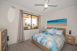 Een slaapkamer met een bed, een nachtkastje, een lamp, een plafondventilator, een raam met gordijnen en een schilderij van een strand.