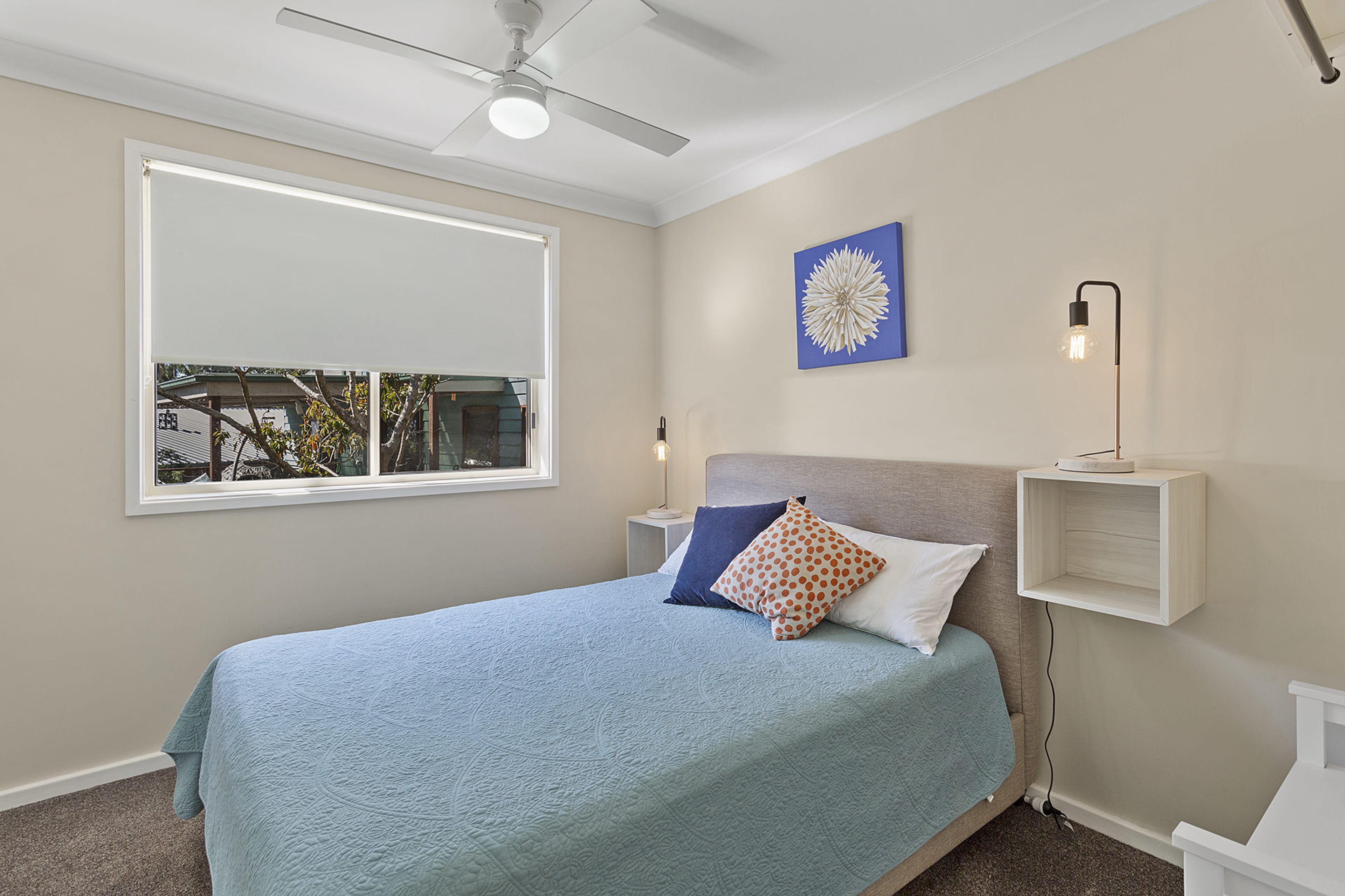 A bedroom with a bed, a window, a ceiling fan, a wall-mounted light, and a decorative wall piece.