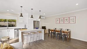 Eine moderne Küche mit weißen Schränken, einem Edelstahl-Kühlschrank und einer Bar mit Hockern. Es gibt einen Esstisch mit Stühlen und gerahmte Kunstwerke an der Wand.