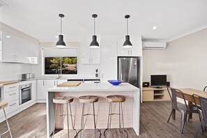 A modern kitchen with white cabinetry, a central island with stools, pendant lighting, and a built-in TV. - Nelson Bay Ocean Jewel (Nelson Bay)
