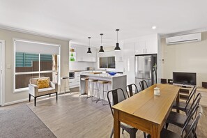 A modern kitchen with white cabinetry, a stainless steel refrigerator, and a dining area with a wooden table and chairs. - Nelson Bay Ocean Jewel (Nelson Bay)