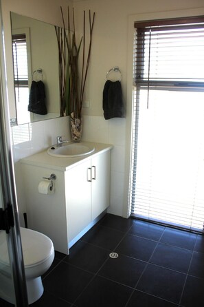 Une salle de bain avec un lavabo blanc, un miroir et des toilettes. Deux serviettes sont accrochées à des crochets, et il y a une fenêtre avec des stores.