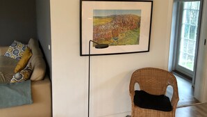 Un salon moderne avec une grande fenêtre, un tableau représentant un paysage encadré, un fauteuil en osier et un canapé agrémenté de coussins à motifs.