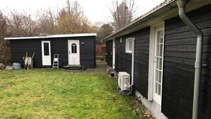 Une maison en bois peinte en noir, avec une porte et des fenêtres blanches, un petit abri de jardin et une cour couverte d’herbe.