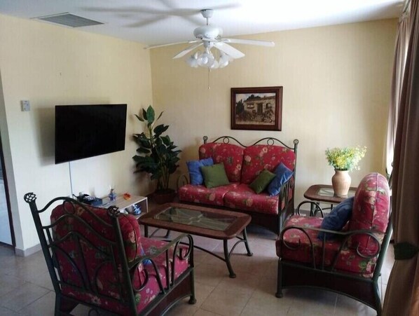 A living room with a ceiling fan, a flat-screen TV, a floral sofa, a coffee table, and a potted plant.