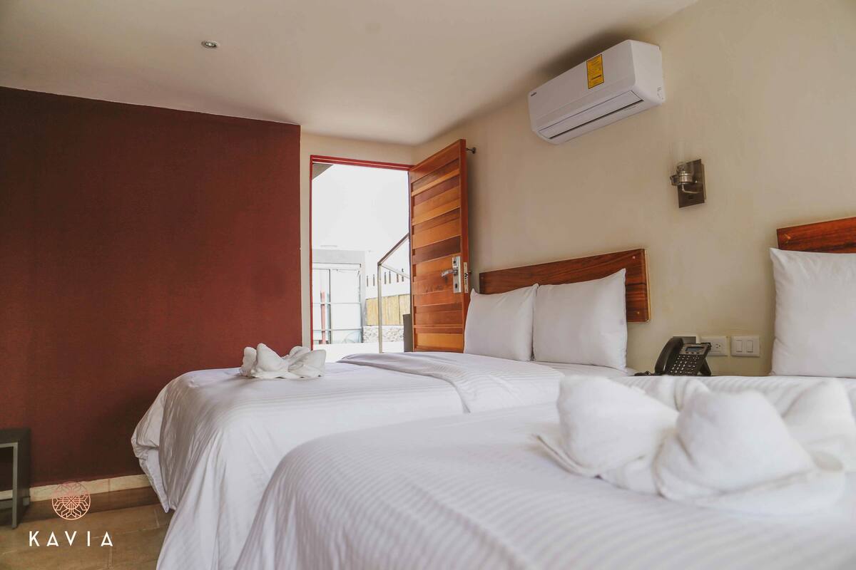 A hotel room with two beds, white bedding, an air conditioning unit, a bedside phone, and a view of the outside.