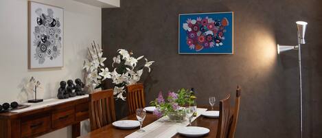 A dining area with a wooden table set for four, surrounded by chairs. There are two framed artworks on the wall, a vase with white flowers, and a floor lamp.