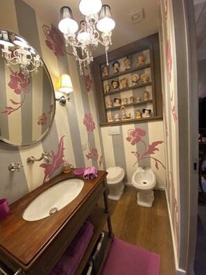 A bathroom with a wooden vanity, a round mirror, a chandelier, a shelf with decorative items, a toilet, and a bidet.