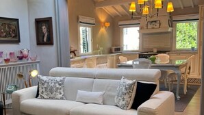 A living room with a sofa, a coffee table, and a dining area with a table and chairs. There is a kitchen visible in the background with a window and a hanging light fixture. - Florence Loft Duomo (Firenze)