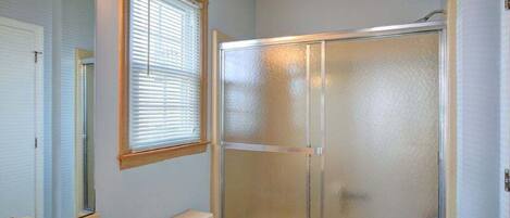 A bathroom with a glass shower enclosure, a sink with a mirror, and a window with blinds.