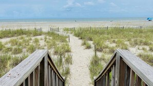 Un sentier bordant la plage et menant à l’océan, avec une clôture en bois et de l’herbe au premier plan.