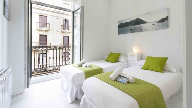 Habitación de hotel con dos camas, un balcón que ofrece vistas a edificios, un cuadro en la pared y una mesita de noche con lámpara.