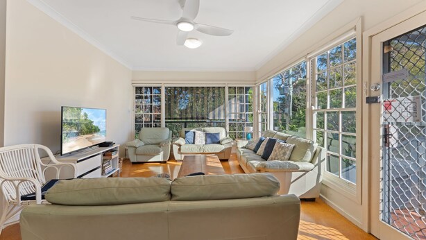 A spacious living room with a large sofa, armchairs, a coffee table, and a TV. There are large windows with a view of trees and a ceiling fan.