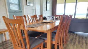 Une table à manger en bois, accompagnée de six chaises, un set de table en fibres tressées et une vue sur la mer grâce à de grandes fenêtres.