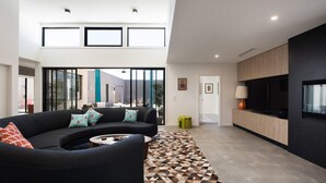 Ein modernes Wohnzimmer mit einer schwarzen Couch, gemusterten Kissen, einem Couchtisch und einem Teppich mit geometrischem Muster. Ein großes Fenster bietet Blick ins Freie, ein Küchenbereich mit einem schwarzen Fernseher und eine Tür zu einem weiteren Raum.