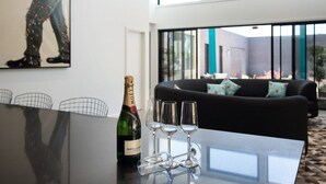 Ein modernes Wohnzimmer mit einem schwarzen Sofa, einem Glastisch mit Champagnerflasche und Gläsern sowie einem großen Fenster mit Blick ins Freie.