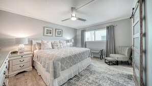 Ein Schlafzimmer mit einem Bett, Nachttischen, einem Schreibtisch, einem Sessel, einem Deckenventilator und einem Fenster mit Vorhängen.