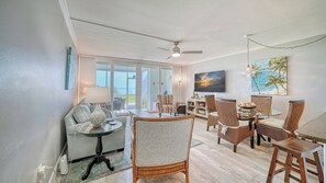 A spacious living area with a glass door leading to a balcony, wicker furniture, and a painting of a sunset.