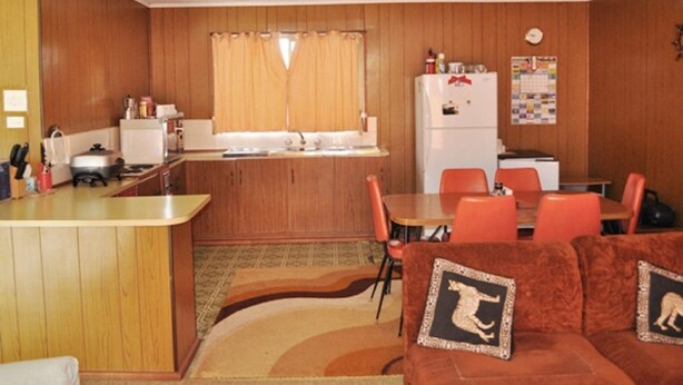 A compact kitchen with wooden paneling, a refrigerator, a sink, and a dining area with orange chairs and a brown sofa.