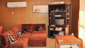 A living room with a brown sectional sofa, a wooden coffee table, a TV on a wooden stand, and a bookshelf.