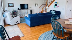 Un salon avec un canapé bleu, un fauteuil blanc, un téléviseur, une table à manger en bois et un escalier.