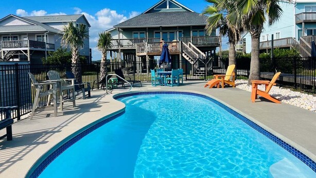 Une piscine avec des chaises longues, une terrasse couverte et un bâtiment à deux étages doté de balcons.