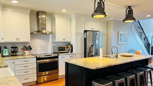 Une cuisine moderne avec des meubles blancs, des appareils en acier inoxydable, un grand îlot central doté de tabourets et un escalier menant à un autre niveau.