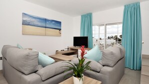 Ein modernes Wohnzimmer mit einem grauen Sofa, einem Couchtisch mit Pflanze, einem Flachbildfernseher und einem Gemälde mit Strandmotiv.