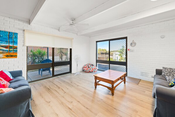 A spacious living area with a wooden floor, a ceiling fan, and a large window with a view of the outdoors. - Lentara Pet Friendly Fingal Bay Home (Fingal Bay)