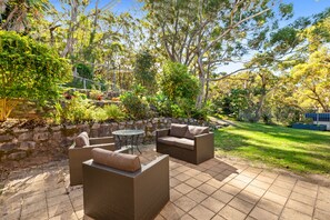 An outdoor patio with wicker furniture, a stone wall, and a grassy area. - Lentara Pet Friendly Fingal Bay Home (Fingal Bay)