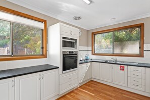 A modern kitchen with white cabinetry, a black countertop, and a view of trees through the windows. - Lentara Pet Friendly Fingal Bay Home (Fingal Bay)