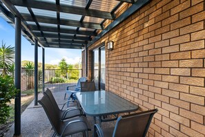 Een terras buiten met een tafel, stoelen, een bakstenen muur en een overkapping van metaal.