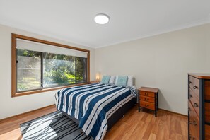 A bedroom with a large window, a bed with striped bedding, a nightstand, a dresser, and a ceiling light. - Lentara Pet Friendly Fingal Bay Home (Fingal Bay)