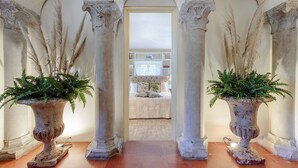 Un pasillo con tres columnas, cada una coronada con una planta. Al final hay una puerta que da a una habitación con una cama y una estantería.