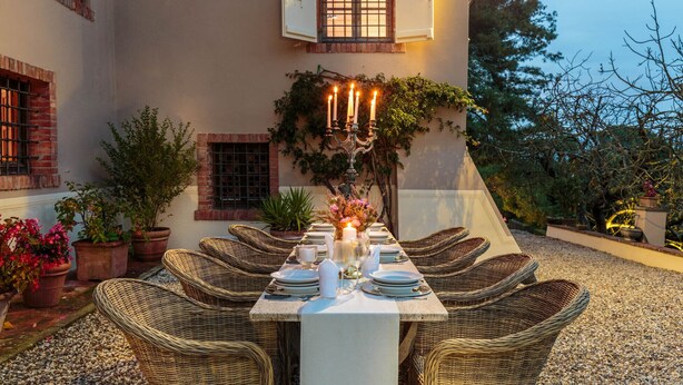 Un área de comedor al aire libre con una mesa dispuesta para seis personas, rodeada de sillas de mimbre, frente a un edificio con fachada de ladrillo y una ventana con persianas.