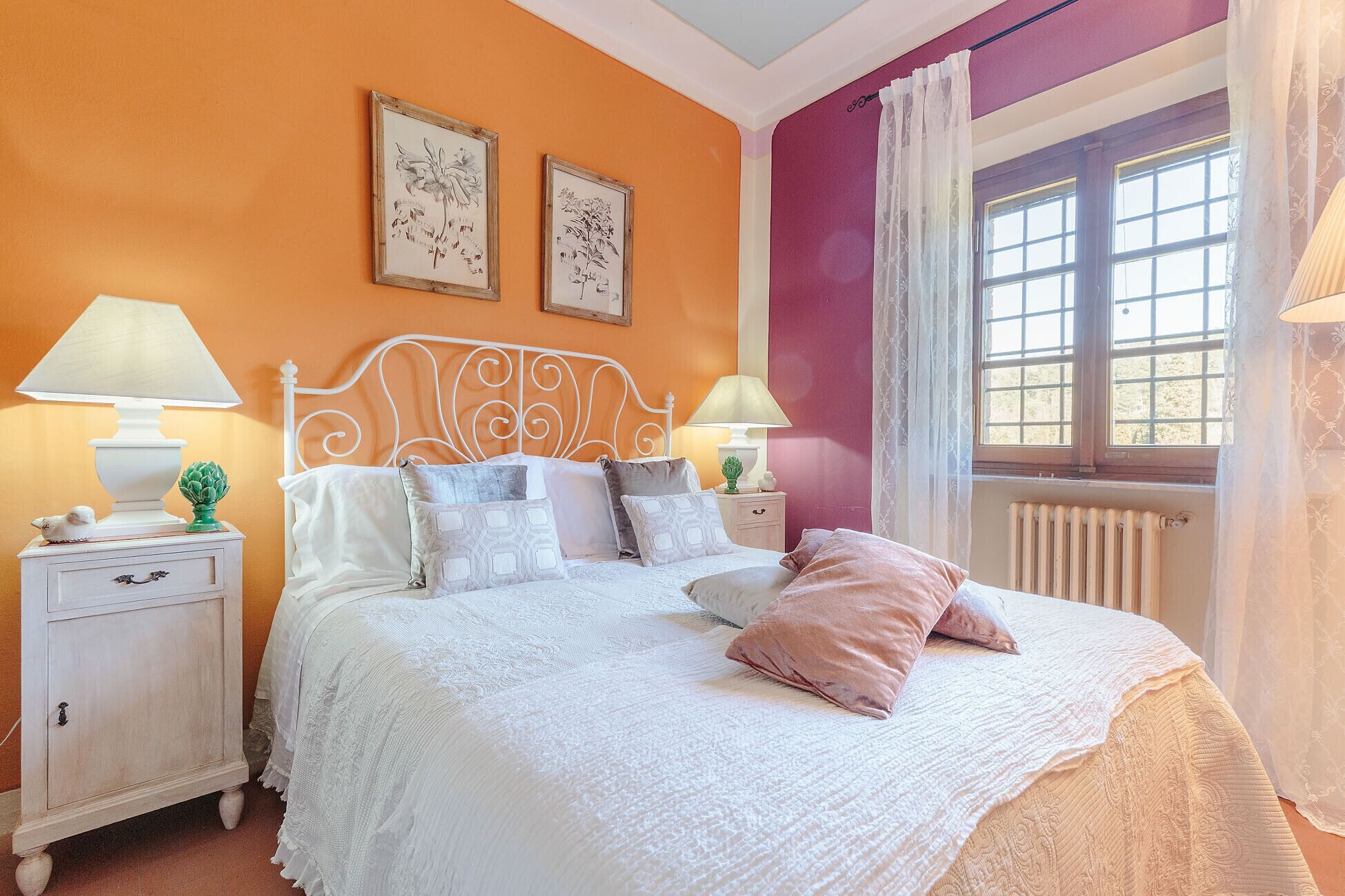 Een slaapkamer met een wit bed, twee nachtkastjes, een raam met gordijnen, en twee ingelijste botanische prints aan de muur.