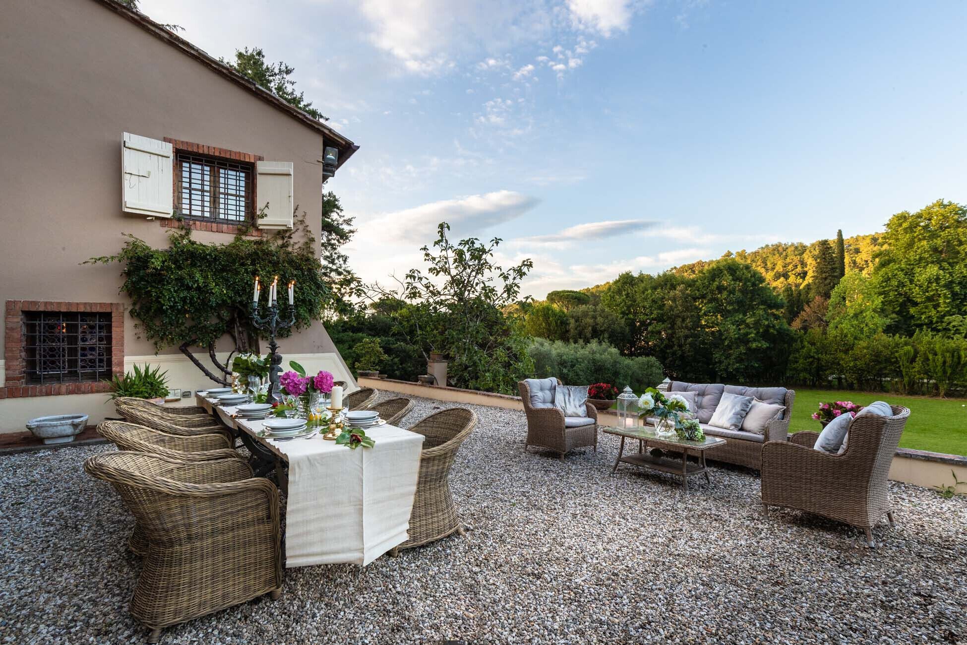 An outdoor dining area with wicker furniture, a table set for four, and a backdrop of a house and lush greenery.