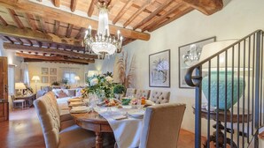 Un comedor con una mesa de madera dispuesta para una comida, rodeada de sillas tapizadas. Un candelabro cuelga del techo, y hay obras de arte enmarcadas en las paredes.