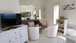 A modern living room with a flat-screen TV, white furniture, a large mirror, and a view of greenery outside. - Bright seafront house with superb sea view, 2 free parking spaces. (Cap d'Agde)