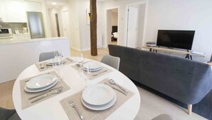 A modern living room with a round dining table set for four, a grey sofa, a flat-screen TV, and a kitchen area with white cabinets and a refrigerator. - Verona | The Rentals Collection (San Sebastián/Donostia)