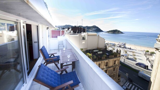 A balcony with blue cushioned chairs and a table, overlooking a beach and a cityscape. - Niza | The Rentals Collection (San Sebastián)
