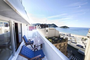 Un balcon avec des fauteuils bleus dotés de coussins et une table, d’où l’on peut admirer une plage et le paysage urbain.
