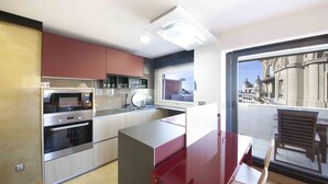 A modern kitchen with stainless steel appliances, a red dining area, and a view of a historic building through a large window. - Niza | The Rentals Collection (San Sebastián)