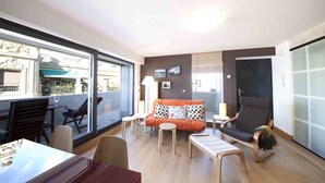 A modern living room with a sofa, chairs, a table, and a large window offering a view of the outside. - Niza | The Rentals Collection (San Sebastián)