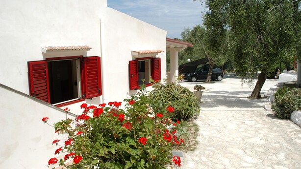 A white building with red shutters, a stone pathway, and red flowers in the foreground.