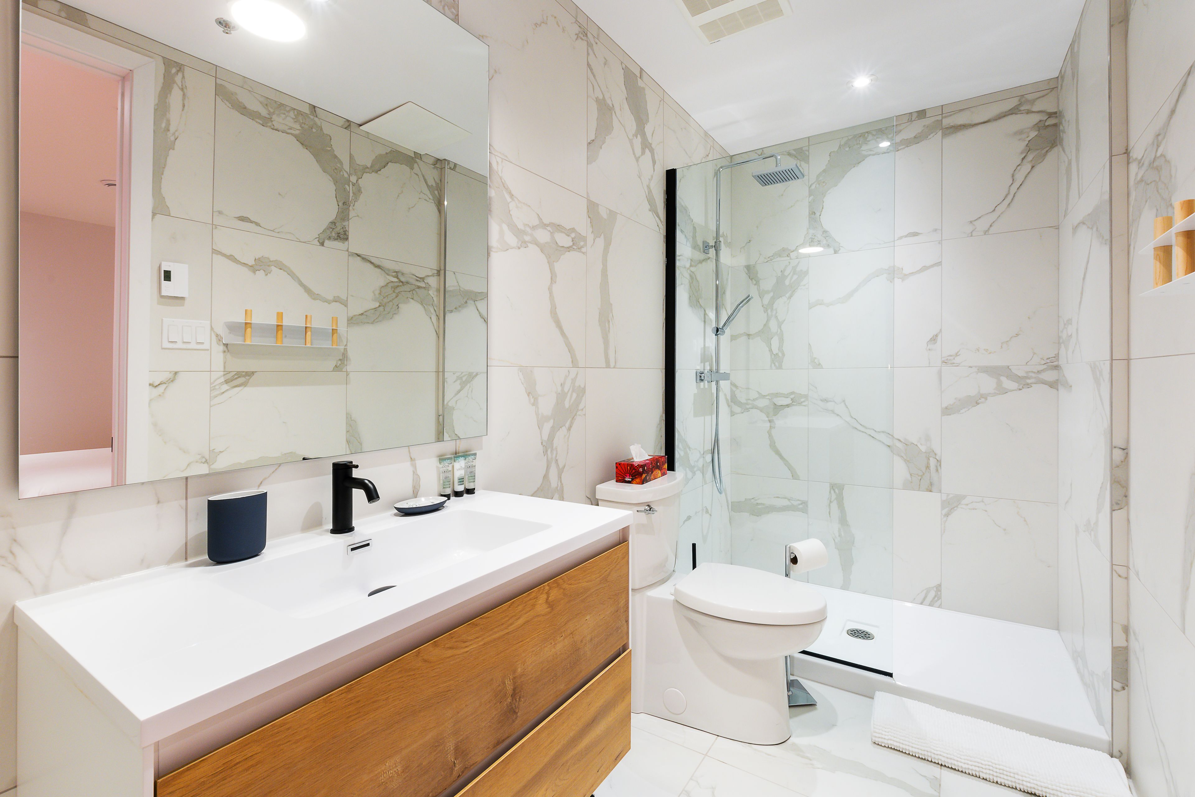 A modern bathroom with a marble backsplash, a white sink, a wooden vanity, and a glass shower enclosure.
