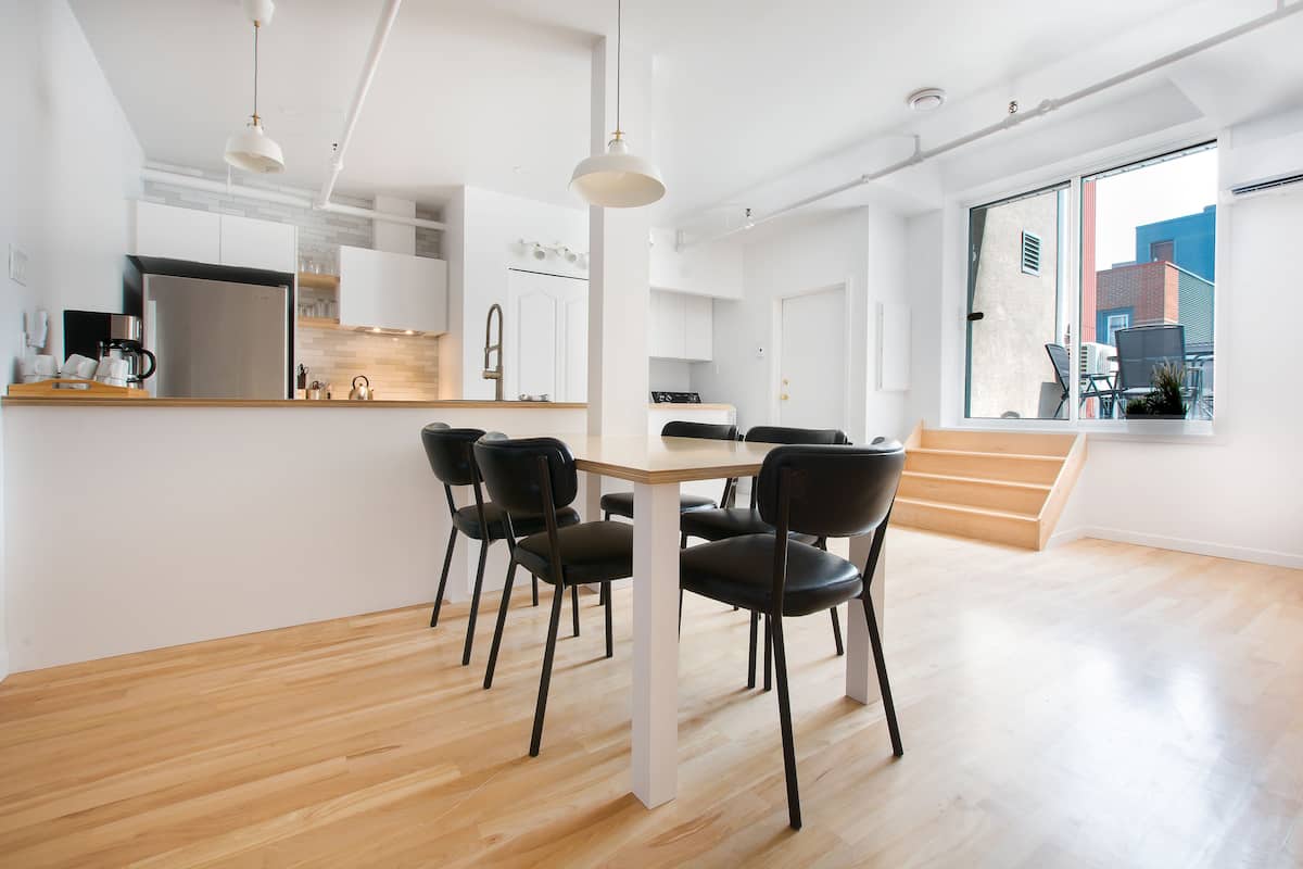 Une cuisine moderne avec des armoires blanches, un réfrigérateur en acier inoxydable et un plan de travail en bois. On y trouve un coin repas avec une table et des chaises, ainsi qu’une grande fenêtre offrant une vue sur la ville.