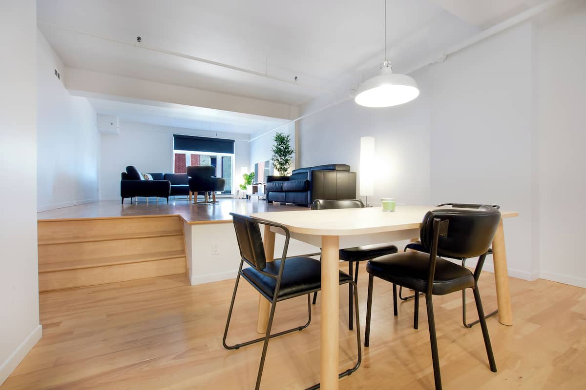 Un espace de repas moderne avec une table en bois et des chaises noires, un escalier menant à une autre pièce et une vue sur le paysage urbain par le biais d’une grande fenêtre.