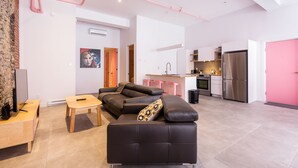 Un salon moderne avec un canapé d’angle, une table basse et un meuble pour la télévision. On y trouve également un coin cuisine avec un réfrigérateur et un mur rose.