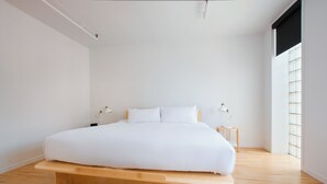 Une chambre à coucher au style minimaliste, avec un lit sur une plateforme en bois, du linge de lit blanc, deux tables de chevet avec des lampes et une grande fenêtre avec des stores.